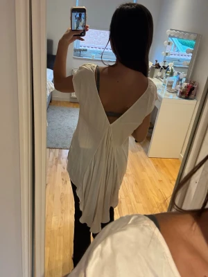 Vit topp med öppen rygg och drapering - Säljer en vit topp med lös passform och korta ärmar. Toppen har en djup v-ringning i ryggen och snygg drapering baktill som ger en unik look. Materialet ser ut att vara mjukt och faller fint. Perfekt för dig som vill sticka ut lite extra.
