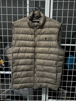 Beige dunväst från Morris Stockholm - Snygg beige dunväst från Morris Stockholm med diskret logga på bröstet. Västen har hög krage, dragkedja framtill och quiltad design. Perfekt för lager på lager och ger en clean look till din outfit.