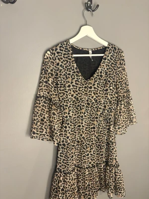 Leopardmönstrad kortklänning med volang - Säljer en snygg kort klänning med leopardmönster och v-ringning. Klänningen har trekvartsärm och volangdetalj nedtill, vilket ger en trendig och avslappnad look. Perfekt för dig som vill sticka ut lite extra.
