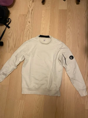 Vit sweatshirt från C.P. Company - Snygg vit sweatshirt från C.P. Company med rund halsringning och ikonisk svart linsdetalj på ärmen. Tröjan har en ficka med dragkedja på vänster ärm och ribbade muddar vid ärmslut och nederkant. Perfekt för en clean och stilren look.