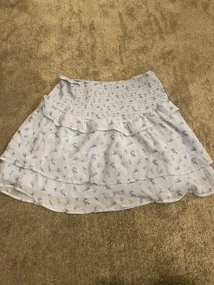 Ljusblå blommig volangkjol - Supersöt kort kjol i ljusblått med små blommor och volangdetaljer. Kjolen har smock i midjan för skön passform och är gjord i ett lätt, luftigt material. Perfekt för varma dagar och ger en romantisk vibe till din outfit.