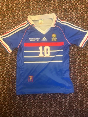 Frankrike VM 98 matchtröja Adidas Zidane - Klassisk blå Frankrike matchtröja från VM 98 med nummer 10 på bröstet och även där bak zidane. Tröjan har vita och röda detaljer, V-ringad krage och broderade loggor. Tillverkad i lätt och ventilerande material, perfekt för fotboll eller samling.