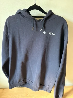 Mörkblå hoodie med tryck Ma Chérie - Mysig mörkblå hoodie med huva och dragsnören. På bröstet står det 'MA CHÉRIE' i vitt och på ryggen finns ett coolt tryck med hjärtan och pilar där de också står ’MA CHÉRIE’.Hoodien har ribbade muddar vid ärmar och nederkant. Perfekt för en avslappnad och trendig look.