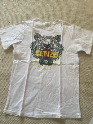 Vit Kenzo t-shirt med tigertryck - Säljer en vit t-shirt från Kenzo med det ikoniska tigertrycket och färgglad logga på bröstet. T-shirten har korta ärmar och är gjord i mjuk bomull. Perfekt för dig som vill sticka ut med en klassisk streetwear-look. Står XL på tröjan men sitter som en S.