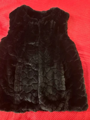 Svart fluffig fuskpäls väst S  - Snygg svart pälsväst i mjukt fuskpälsmaterial med ett coolt mönster i pälsen. Västen har en rak passform och är ärmlös, perfekt att slänga över en hoodie eller tröja för extra stilpoäng. Stängs framtill och har en mysig känsla.