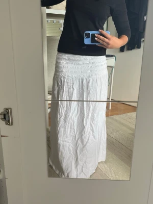 Vit långkjol med smock i midjan - Säljer en enkel och snygg vit långkjol med bred smock i midjan. Kjolen är rak i modellen och gjord i ett lätt och luftigt material, perfekt för varma dagar. Passar till både sneakers och sandaler för en chill look.