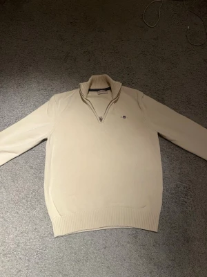 Beige stickad tröja med halv dragkedja - Snygg beige stickad tröja med halv dragkedja och hög krage. Tröjan har långa ärmar, ribbade muddar och en liten broderad logga på bröstet. Perfekt för lager på lager och enkel att matcha med olika outfits.väldigt bra skick säljer för 550 kan gå med på priser ner till 400