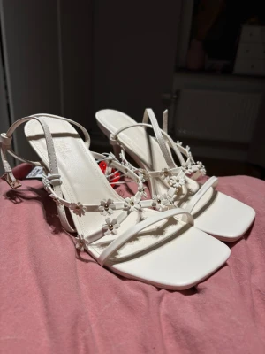 Vita öppna sandalletter  - Somriga vita sandalletter med blomdetaljer och justerbara remmar från NEW YORKER. I storlek 40 men upplevs som små i storleken. Klacken är 3cm. Helt oanvända med prislapp kvar. Köptes för 299kr. Materialet är konstläder.