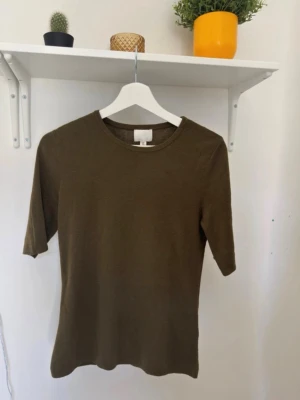 Olivgrön topp från H&M - En enkel och stilren olivgrön topp från H&M med rund halsringning och halvkort ärm. Toppen har en rak passform och är tillverkad i ett mjukt material som känns skönt mot huden. Perfekt att matcha med jeans eller kjol.