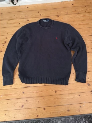 Mörkblå stickad tröja Polo Ralph Lauren - Mörkblå stickad tröja från Polo Ralph Lauren med rund halsringning och ribbade muddar. Klassisk design med röd broderad logga på bröstet. Storlek L men passar mer som M