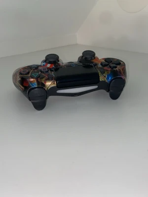 Flerfärgad PS4 handkontroll - Säljer en unik handkontroll till PlayStation 4 med ett färgstarkt och detaljerat mönster över hela ytan. Kontrollern har klassisk form med två styrspakar, touchpad och alla vanliga knappar. Materialet är plast och designen sticker verkligen ut bland vanliga kontroller.