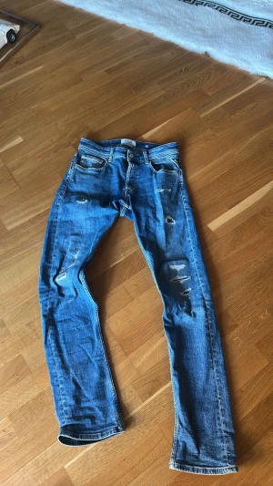 Blå slitna jeans med raka ben - Säljer ett par blå jeans med slitningar och raka ben. Jeansen har klassisk femficksdesign, normal midja och snygga slitdetaljer på både fram- och baksida. Perfekta för en avslappnad och trendig look. 