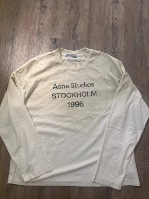 Vit långärmad topp Acne Studios - Vit långärmad topp från Acne Studios med tryck 'Acne Studios STOCKHOLM 1996' på bröstet. Klassisk rund halsringning och relaxed fit. Perfekt till jeans eller kjol för en clean och stilren look.