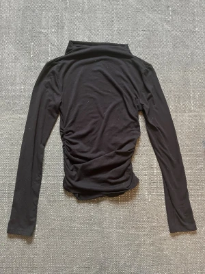 Svart polotopp med rynkade sidor - Säljer en svart polotopp med långa ärmar och snygga rynkade detaljer på sidorna. Toppen har en tight passform och är tillverkad i ett mjukt, stretchigt material som sitter skönt på kroppen. Perfekt att styla med jeans eller kjol. Lite nopprig men inget man tänker på. Från Kappahl i storlek XS.