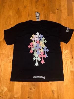 Svart Chrome Hearts T-shirt - Svart t-shirt med flera färgglada kors i pastellfärger på ryggen och ett mindre tryck framtill. T-shirten har rund hals, korta ärmar och är gjord i mjuk bomull. 