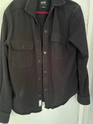 Svart overshirt från Only & Sons - Snygg svart overshirt från Only & Sons i relaxed fit. Skjortjackan har två bröstfickor med lock och knappar, klassisk krage och tryckknappar framtill. Materialet är kraftigt och passar perfekt för lager på lager-stil.