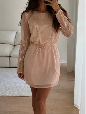 Ljusrosa kortklänning med paljetter från oasis - Supersöt ljusrosa kortklänning från Asos med transparenta långärmar och glittriga paljettdetaljer i blommigt mönster. Klänningen har ett tunt bälte i midjan och rund halsringning. Perfekt för dig som vill sticka ut med en elegant look.