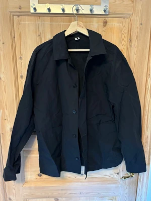 Svart overshirt med knappar från Arket - Stilren svart overshirt med klassisk krage och knäppning framtill. Overshirten har en rak passform och långa ärmar. Perfekt att bära som lager över en t-shirt eller hoodie.                                 Tunt polyester liknande material