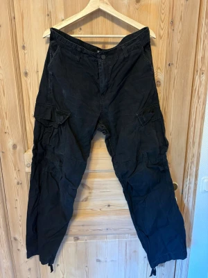 Svarta cargobyxor från Carhartt - Säljer ett par svarta cargobyxor från Carhartt med klassiska cargofickor på sidorna och dragsko vid benslut. Byxorna har rutmönstrad struktur och är tillverkade i slitstarkt bomullstyg. Perfekta för en streetwear-look.
