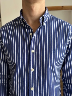 Blåvit randig skjorta från Polo Ralph Lauren (Slim Fit) - Snygg blå skjorta med vita vertikala ränder från Ralph Lauren. Klassisk button-down krage, långa ärmar och broderad logga på bröstet. Skjortan har normal passform och är tillverkad i bomull. Perfekt för en stilren look. Storlek M. Snabb Leverans!