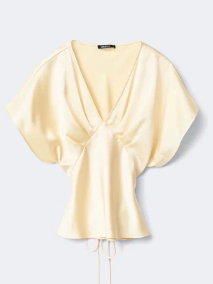 Krämfärgad satintopp med v-ringning - Snygg krämfärgad blus i glansigt satinmaterial med djup v-ringning och korta ärmar. Modellen har en figurnära passform med knytband i midjan som ger en feminin siluett. Perfekt för att lyfta vilken outfit som helst.