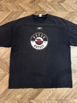 Svart Reebok Petes Hockey t-shirt - Säljer en svart t-shirt från Reebok med Petes Hockey-tryck på bröstet. T-shirten har rund hals och korta ärmar. Tillverkad i 100% bomull, perfekt för dig som gillar sportig stil och hockey.