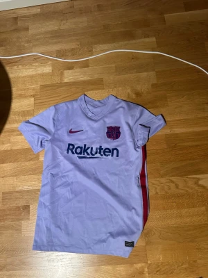 Ljuslila FC Barcelona matchtröja - Säljer en ljuslila FC Barcelona matchtröja med korta ärmar. Tröjan har klubbmärke och Nike-logga på bröstet, Rakuten-tryck framtill och Unicef-tryck bak. Sidorna har röda och blå detaljer. Perfekt för dig som älskar fotboll och Barca!
