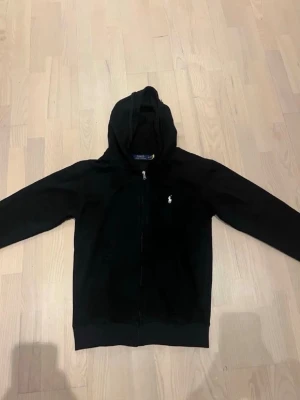 Svart hoodie från Polo Ralph Lauren - Snygg svart hoodie från Polo Ralph Lauren med dragkedja framtill och klassisk huva. Liten vit logga broderad på bröstet. Tillverkad i mjukt bomullsmaterial som är skönt att ha på sig. Perfekt för en chill och stilren look.