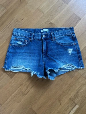 Blå jeansshorts med slitningar - Säljer ett par blå jeansshorts med råa, fransiga kanter och slitna detaljer framtill. Klassisk femficksmodell med bälteshällor och knappgylf. Perfekta för varma dagar och ger en avslappnad vibe. De är lågmidjade.