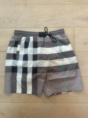 Rutiga shorts med snörning - Snygga shorts med stort rutmönster i svart, vitt och grått samt en diskret grön linje. Shortsen har elastisk midja med snörning och sidofickor. Perfekta för sommaren och ger en trendig vibe till din outfit.