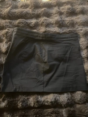 Svart kortkjol med dragkedja - En enkel svart kortkjol med rak passform och diskret dragkedja i sidan. Kjolen är tillverkad i ett slätt tyg och har en klassisk midja. Perfekt att matcha med olika toppar för en clean look.
