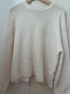 Krämvit oversized stickad tröja - Mysig och oversized stickad tröja i krämvit färg med rund halsringning och ribbade muddar vid ärmslut och nederkant. Perfekt för lager på lager och en avslappnad stil. Materialet ser ut att vara mjukt och fluffigt, troligen ull- eller akrylblandning.