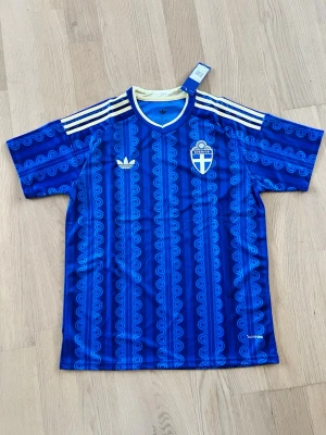 Blå Sverige Adidas fotbollströja - Säljer en blå fotbollströja från Adidas med Sveriges landslagsemblem på bröstet. Tröjan har ett retroinspirerat mönster, vita Adidas-ränder på axlarna och vit krage. Perfekt för match eller träning. Materialet är lätt och andas.