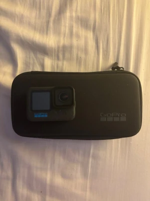 GoPro 12 black - GoPro 12 med tillhörande skyddsväska, batteri, handgrepp med orange detalj, fästen och skruvar samt handledsrem. Allt är kompakt och lätt att ta med sig.