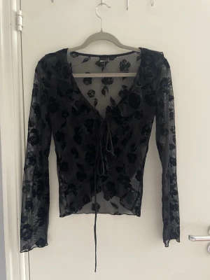 Svart mesh topp med sammetsrosor - Snygg svart mesh topp med långa ärmar och sammetsrosor i mönstret. Djup V-ringning och knytband framtill ger en trendig look. Perfekt att styla lager på lager eller som statement-plagg. Transparent material för en edgy vibe.