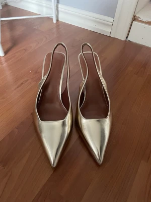 Guldiga slingback pumps med spetsig tå - Snygga pumps i metallic guld med spetsig tå och öppen häl. Skorna har smal klack och elegant slingback-rem. Tillverkade i ett glansigt material som ger en lyxig känsla och passar perfekt till trendiga outfits.