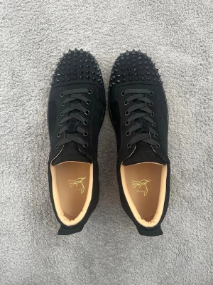 Svarta sneakers med nitar - Säljer ett par svarta sneakers från Christian Louboutin med coola svarta nitar på tån och klassiska röda sulor. Skorna har svarta skosnören och en lyxig känsla med beige innersula. Perfekta för dig som vill sticka ut med din stil. Finns dessutom 2 nitar i en liten påse om nitarna skulle försvinna eller bli skadade.