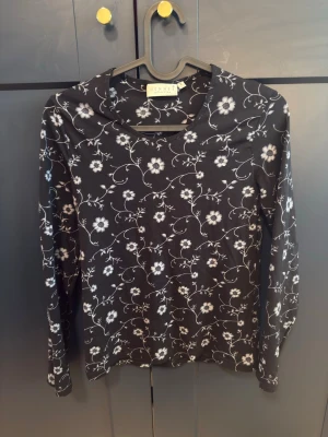 Svart blommig långärmad topp - Svart långärmad topp med vitt blommönster och bred krage. Tunn och mjuk känsla, perfekt för dig som gillar en enkel men ändå söt stil. Passar till både jeans och kjol för en chill look.