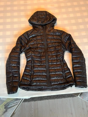 Everest dunjacka  - Snygg svart dunjacka med huva och dragkedja framtill. Jackan har quiltad design med horisontella sömmar och figurnära passform. Perfekt för kalla dagar när du vill hålla värmen och ändå se stilren ut.