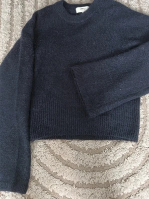 Mörkblå stickad tröja Gina Tricot - Mysig mörkblå stickad tröja från Gina Tricot i ullblandning. Tröjan har rund halsringning, långa ärmar och ribbstickad nederkant. Perfekt för kyliga dagar och enkel att matcha med jeans eller kjol. (Breda och utsvängda armar)