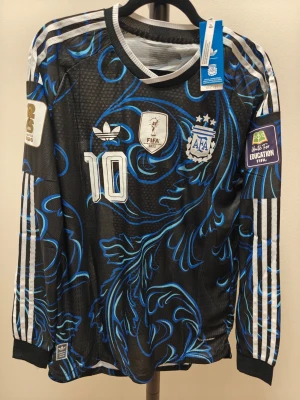 Argentina semifinal Messi långärmad spelar version - Säljer en svart långärmad Argentina matchtröja från Adidas med blått och vitt mönster, nummer 10 och Messi på ryggen. Tröjan har officiella FIFA-märken på ärmarna och tre vita ränder längs axlarna. Perfekt för fotbollsfans som vill sticka ut.