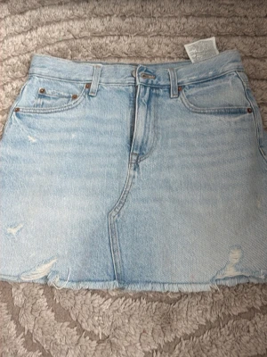 Ljusblå jeanskjol från Zara - Säljer en ljusblå jeanskjol från Zara med rå kant nertill och slitna detaljer. Kjolen har klassisk femficksdesign och bälteshällor. Perfekt för dig som gillar en avslappnad och trendig stil.