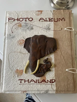 Vintage Fotoalbum med elefanter Thailand - Unikt fotoalbum med handgjort omslag i beige och brunt papper, dekorerat med upphöjda elefanthuvuden i brunt och elfenbensfärg. Texten 'PHOTO ALBUM' och 'THAILAND' på framsidan. Knytband på sidan för stängning. Perfekt för minnen från resor.