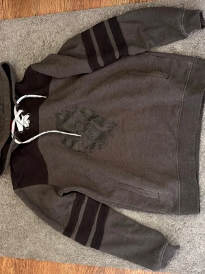 Grå hoodie med Modo Hockey-tryck - Snygg grå hoodie med svart Modo Hockey-logga på bröstet. Tröjan har svarta detaljer på axlar och ärmar samt vita snören i huvan. Perfekt för dig som gillar sportig stil och vill visa ditt stöd för Modo Hockey.
