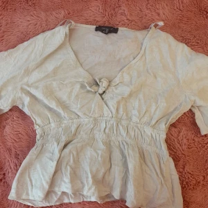 Vit blus med knyt och volang - Söt vit blus med v-ringning, knyt framtill och volang nedtill. Blusen har korta ärmar och resår i midjan för en feminin siluett. Perfekt till jeans eller kjol för en fräsch look.