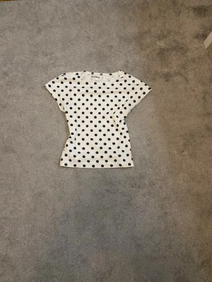 Vit prickig topp med kort ärm - Snygg vit topp med svarta prickar och korta ärmar. Toppens passform är figurnära och materialet känns mjukt och stretchigt. Perfekt för dig som gillar lekfulla mönster och en stilren look.