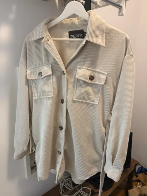 Beige manchester overshirt från Pieces - Säljer en beige overshirt från Pieces i mjuk manchester. Skjortan har långa ärmar, två stora bröstfickor med knappar och klassisk krage. Perfekt att slänga över en t-shirt för en chill look.