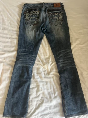 Low rise blå jeans från Z-Brand - Säljer ett par blå jeans med slitningar och unika broderade bakfickor. Byxbenen har dragkedjor som ger ett coolt intryck. Jeansen har raka ben med lite utsvävning och klassisk passform, perfekta för dig som gillar detaljer. Jag är en storlek 38 och dom passar mig bra i midjan💓