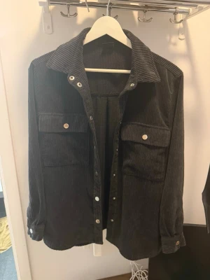 Svart manchester overshirt Gina Tricot - Säljer en svart overshirt i manchestertyg från Gina Tricot. Skjortan har två stora bröstfickor med knappar, klassisk krage och tryckknappar framtill. Perfekt lager på lager-plagg med avslappnad passform och trendig look.