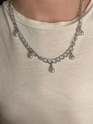 Silverfärgat halsband med pärldroppar - Säljer ett chunky halsband i silverfärgad metall med fem droppformade vita pärlor som hänger längs kedjan. Kedjan har stora länkar och ger ett trendigt uttryck. Perfekt för dig som gillar statement-smycken och vill addera något extra till din outfit.